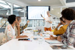 four people sitting at a table with a lady presenting at a whiteboard
